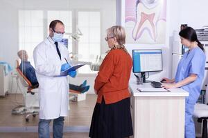 dentiste médecin avec visage masque expliquant diagnostic à Sénior femme patient dans stomatologie attendre zone couloir. personnes âgées homme séance sur chaise pour les dents traitement. photo