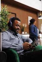 souriant africain américain homme homme d'affaire dans écouteurs en utilisant numérique tablette vérification email pendant affaires voyage, noir gars Hôtel client navigation l'Internet tandis que attendre pour enregistrement dans hall photo