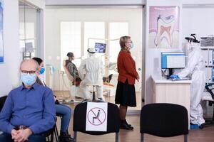 Sénior patient avec visage msak discuter avec dentiste assistant habillé ppe costume en gardant social distanciation dans attendre zone. dentisterie Personnel dans protection costume pendant global pandémie avec COVID-19 [feminine. photo