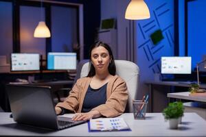 portrait femme d'affaires à la recherche à caméra tandis que travail sur entreprise date limite. intelligent femme séance à sa lieu de travail dans le cours de en retard nuit heures Faire sa emploi. photo