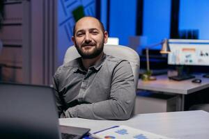 sur de soi entrepreneur à la recherche sur de soi à la recherche à caméra dans vide bureau. intelligent homme d'affaire séance à le sien lieu de travail dans le cours de en retard nuit heures Faire le sien emploi. photo