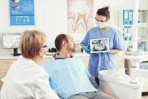 dentiste femme dans dentaire Bureau discuter avec malade homme tandis que infirmière montrant dent radiographie image sur tablette. homme patient attendre mal aux dents traitement séance sur dentaire chaise pendant médical examen photo