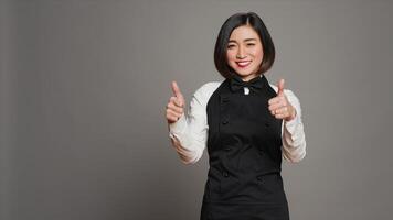 souriant serveuse donnant les pouces en haut signe dans de face de caméra, travail dans une cinq étoile restaurant à servir les tables. asiatique serveur avec tablier montrant approbation avec d'accord symbole dans studio, accord. caméra un. photo