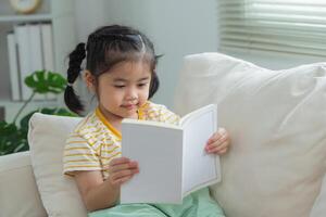 joyeux content asiatique enfant bébé fille souriant et en train de lire livre tandis que séance sur canapé canapé dans vivant pièce à maison. fille relex en train de lire livre sourire à canapé dans le maison. retour à école concept. photo