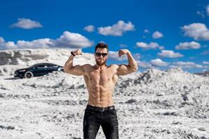 à moitié nu bodybuilder posant avec musclé bras en haut. Contexte de magnifique blanc montagnes ciel et foncé moderne auto. homme dans lunettes de soleil debout. photo