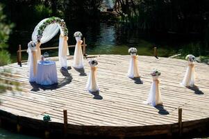 mariage cambre magnifiquement décoré et une petit tableau, bouquets de fleurs préparé pour la cérémonie en plein air. en bois surface dans le forme de une cercle plus de le rivière pour mariage événement. photo