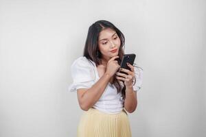 une réfléchi asiatique femme est en portant sa téléphone intelligent tandis que imaginer sa pensées, isolé par blanc Contexte. photo