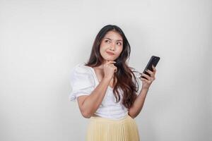 une réfléchi asiatique femme est en portant sa téléphone intelligent tandis que imaginer sa pensées, isolé par blanc Contexte. photo