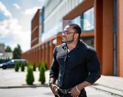 Beau homme avec mains sur le sien taille permanent à le bâtiment. africain homme dans foncé chemise permanent demi-tourné et à la recherche tout droit devant. sportif gars à le affaires centre. photo