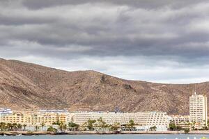 côtier paysage urbain avec hôtels contre montagnes en dessous de nuageux ciels dans los chrétiens, tenerife. photo