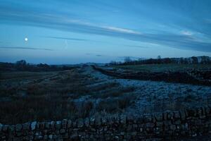 crépuscule plus de une tranquille rural paysage avec une pierre mur et lune visible dans le bleuté ciel. photo