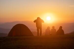 groupe de copains séance par le tente pendant pendant la nuit camping tandis que à la recherche à le magnifique vue point le coucher du soleil plus de le Montagne pour Extérieur aventure vacances Voyage concept photo