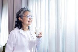 Sénior asiatique Dame en buvant verre de Lait tandis que permanent par le fenêtre avec copie espace pour calcium stimulant et en bonne santé laitier produit consommation photo
