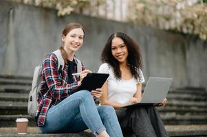 Jeune Université élèves se concentrer sur le sien école projet, à la recherche à portable et tablette, discuter et travail ensemble à le Campus parc photo