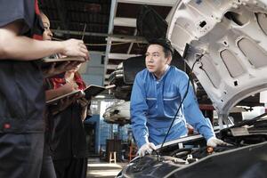 asiatique Masculin professionnel automobile ingénieur superviseur décrit voiture moteur entretien et réparation travail avec mécanicien ouvrier états-majors équipe dans réparer un service garage, spécialiste métiers dans auto industrie. photo