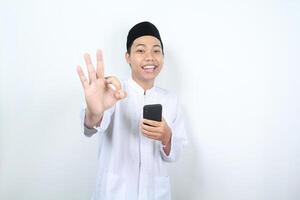 souriant asiatique musulman homme montrant d'accord panneaux avec en portant mobile téléphone isolé photo
