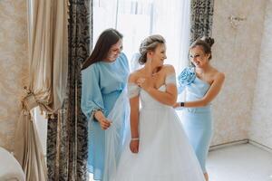 mariage Matin. demoiselles d'honneur Aidez-moi mettre sur le blanc mariage robe. une Jeune femme est en train de préparer à rencontrer sa jeune marié et ayant amusement avec sa copains photo