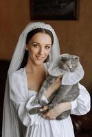 jeune, élégant, fille dans le image de une la mariée avec une magnifique agrafe et une tiare sur sa diriger, en portant sa animal de compagnie chat dans sa bras photo