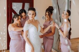 mariage Matin. demoiselles d'honneur Aidez-moi mettre sur le blanc mariage robe. une Jeune femme est en train de préparer à rencontrer sa jeune marié et ayant amusement avec sa copains photo