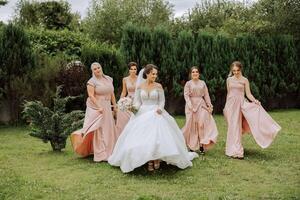 une brunette la mariée et sa demoiselles d'honneur dans identique rose Robes sont en marchant contre le Contexte de la nature. les filles dans identique Robes sont fabrication en dehors à une mariage. mariage dans la nature. photo