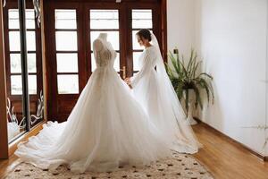 une magnifique la mariée avec une longue voile dans sa chambre, portant une peignoir. mariage robe sur une mannequin. le la mariée dans le Matin avant le mariage cérémonie. photo