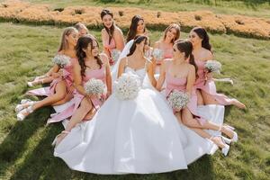 une brunette la mariée et sa demoiselles d'honneur dans identique Robes asseoir avec le la mariée et réjouir avec fleurs dans leur mains sur le vert herbe. mariage dans la nature. photo