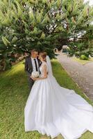 une Beau jeune marié embrasse le sien la mariée dans une luxuriant blanc robe et sourit dans une magnifique Extérieur paramètre. en dessous de le ouvert ciel. haute qualité photo. une nouveau marié couple pose ensemble sur une ensoleillé été journée. photo