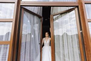 le la mariée des stands par le fenêtre dans une blanc robe et regards à le jeune marié. mariage journée. magnifique la mariée. mariage. photo