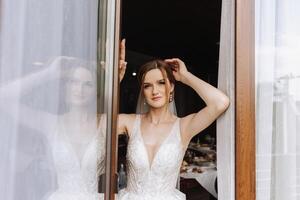 le la mariée des stands par le fenêtre dans une blanc robe et regards à le jeune marié. mariage journée. magnifique la mariée. mariage. photo