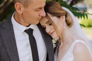 une Beau jeune marié embrasse le sien la mariée dans une luxuriant blanc robe et sourit dans une magnifique Extérieur paramètre. en dessous de le ouvert ciel. haute qualité photo. une nouveau marié couple pose ensemble sur une ensoleillé été journée. photo