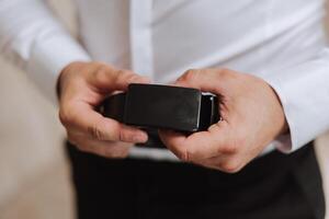 fermer de une homme en portant une noir cuir ceinture. une homme dans une blanc chemise, sans pour autant une affronter, détient une noir cuir ceinture dans le sien mains. le Matin de le jeune marié à le mariage. photo