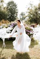 une magnifique brunette la mariée dans un élégant peignoir est en marchant dans une parc avec vert herbe et rose hortensia fleurs. matin, réunions, mariage portrait de une mignonne fille. la photographie et concept. été. photo