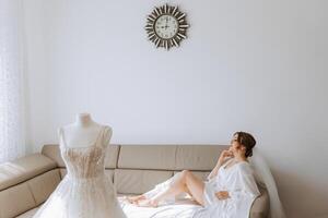 une magnifique la mariée est séance dans une pansement robe dans le Matin avant le mariage la cérémonie dans une Hôtel avec une moderne intérieur. incroyable coiffure de le la mariée. Naturel et moderne se maquiller. photo