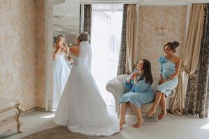 mariage Matin. demoiselles d'honneur Aidez-moi mettre sur le blanc mariage robe. une Jeune femme est en train de préparer à rencontrer sa jeune marié et ayant amusement avec sa copains photo