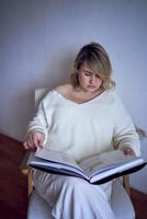 une de taille moyenne femme dans lumière vêtements lit une livre tandis que séance dans une blanc chaise dans une lumière pièce photo