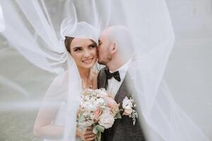 fermer portrait de deux gens dans l'amour. un affectueux jeune marié embrasse le mariée, justificatif sa en dessous de le voile. le meilleur des moments de le mariage. photo