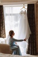 une magnifique la mariée avec une longue voile dans sa chambre, portant une peignoir. mariage robe sur une mannequin. le la mariée dans le Matin avant le mariage cérémonie. photo