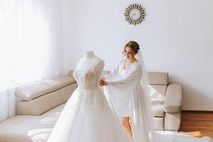 une magnifique la mariée avec une longue voile dans sa chambre, portant une peignoir. mariage robe sur une mannequin. le la mariée dans le Matin avant le mariage cérémonie. photo