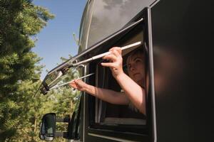 le fille ouvre le fenêtre de le van à prendre plaisir le Soleil. le concept de gens en voyageant sur été vacances à l'intérieur une camping-car, mobile Accueil photo