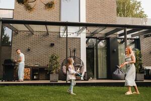 une content famille est repos près leur maison. papa est cuisine une barbecue, et maman et fille sont en jouant photo