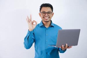 souriant ou content asiatique homme avec des lunettes faire des gestes main D'accord ou terminé et en portant portable portant bleu chemise isolé sur blanc Contexte photo