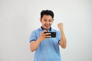 excité ou content Jeune asiatique homme gagner en jouant Jeu avec téléphone intelligent et avec serré les poings isolé sur blanc Contexte photo