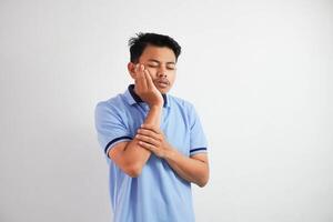 dent mal. Jeune asiatique homme sentiment douleur, en portant le sien joue avec tous les deux mains, Souffrance de mal mal aux dents, ayant douloureux expression portant bleu t chemise isolé sur blanc Contexte photo