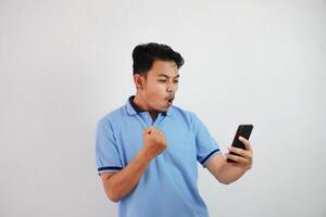 excité ou content portrait Jeune asiatique homme en portant téléphone avec serré les poings portant bleu polo t chemise isolé sur blanc Contexte photo