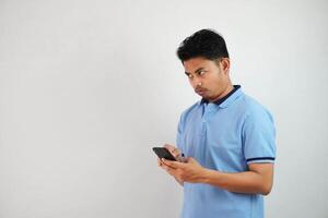 Jeune asiatique homme permanent en portant une téléphone avec un en colère et désapprobateur expression portant bleu t chemise isolé sur blanc Contexte photo