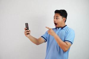 portrait asiatique homme sous le choc en portant téléphone et montrer du doigt à le téléphone avec une doigt portant bleu polo t chemise isolé sur blanc Contexte photo