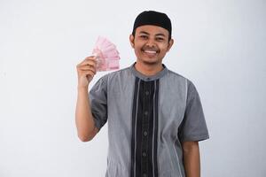 souriant ou content Jeune asiatique musulman homme en portant papier argent cadeau thr de famille Ramadan eid al-fitr temps portant gris musulman vêtements isolé sur blanc Contexte photo