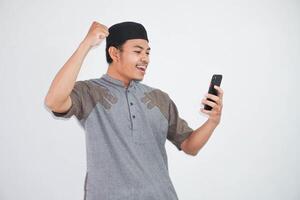 excité ou content Jeune asiatique musulman homme en portant sur mobile téléphone et mains dans les poings Faire gagnant geste portant koko vêtements isolé plus de blanc Contexte photo