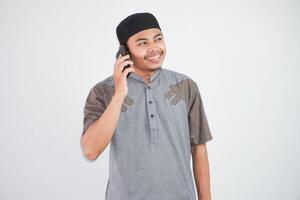 Beau musulman asiatique homme sourire tandis que appel sur une téléphone intelligent portant gris musulman vêtements isolé sur blanc Contexte photo
