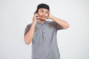 confus Jeune asiatique musulman homme parlant sur mobile téléphone avec mains en portant tête portant gris musulman vêtements isolé sur blanc Contexte photo